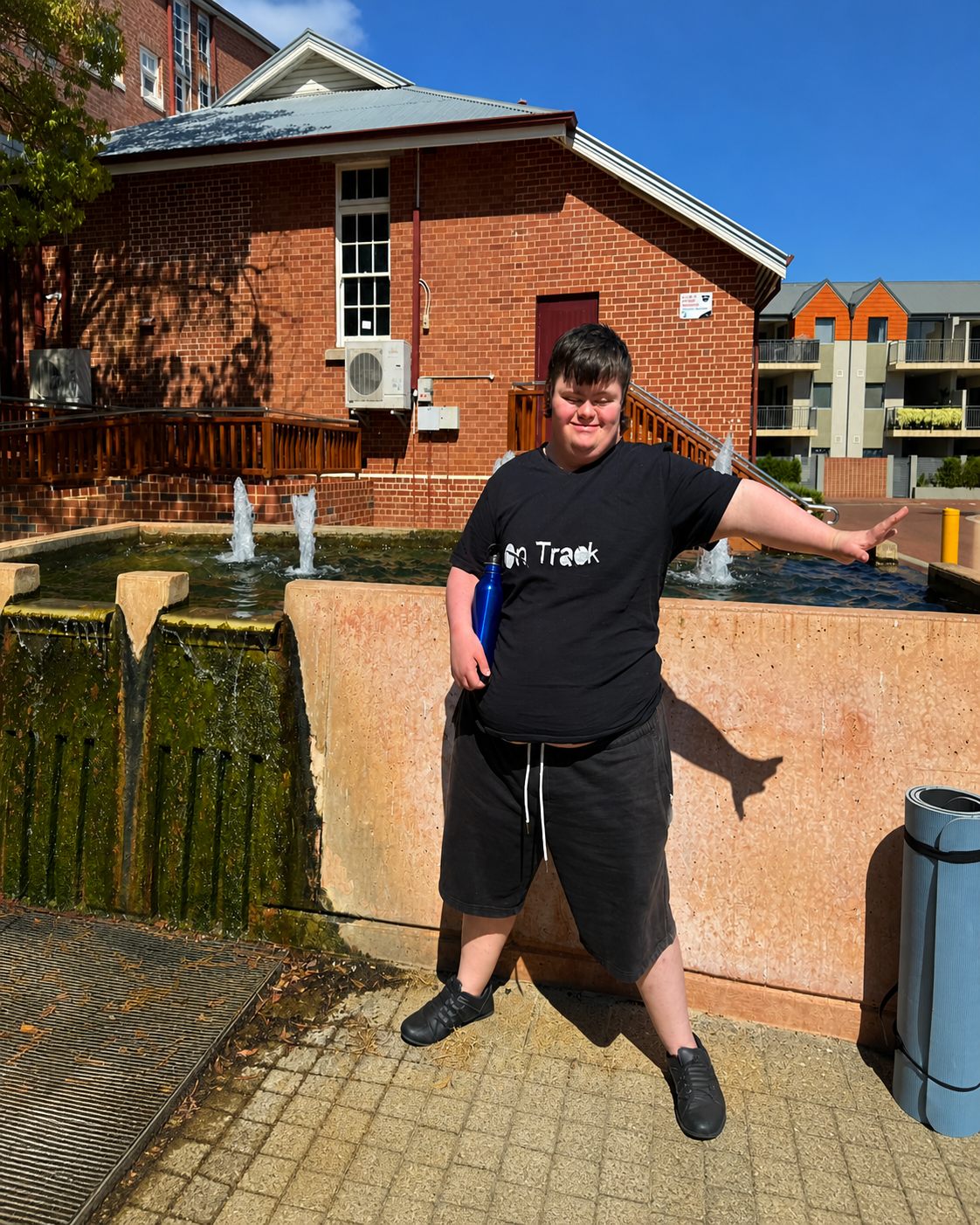 A young man holds a water bottle and strikes a playful pose beside a fountain on a sunny day