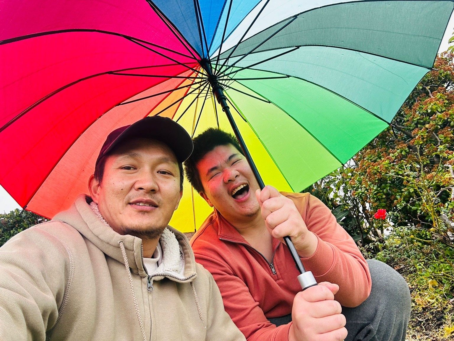 A support worker and a young man share a laugh under a bright rainbow umbrella while sitting in a garden